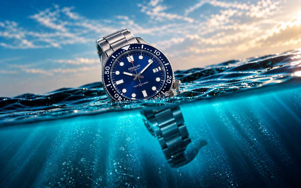 A silver metal wristwatch with a blue dial and blue bezel sits partially submerged at the waterline, with ocean water and a cloudy sky visible above and below the surface.
