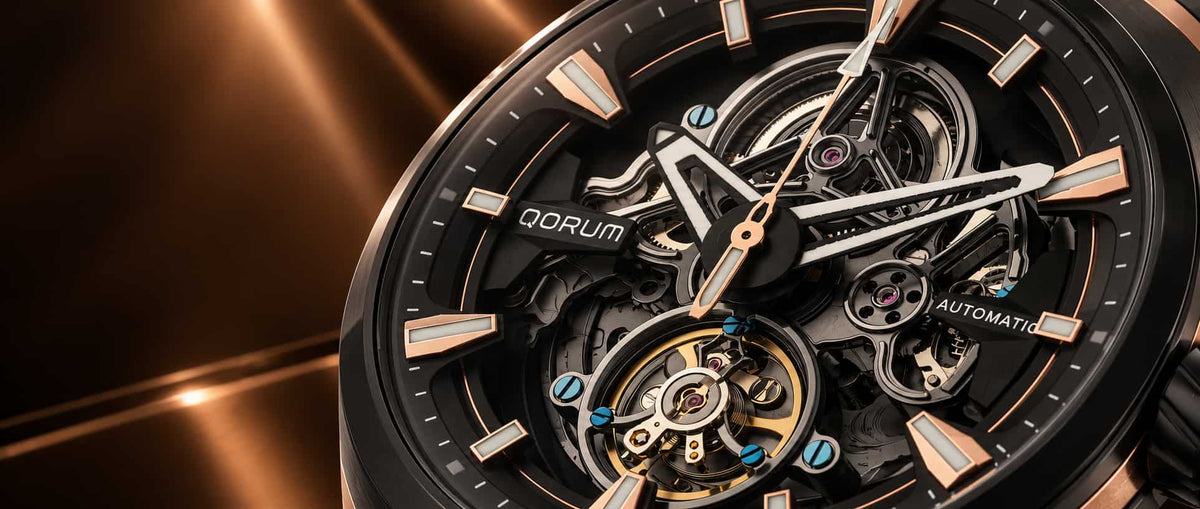 Close-up of a QORUM automatic watch with a black skeleton dial showing intricate internal gears and a gold-tone tourbillon, set against a dark metallic bronze background.