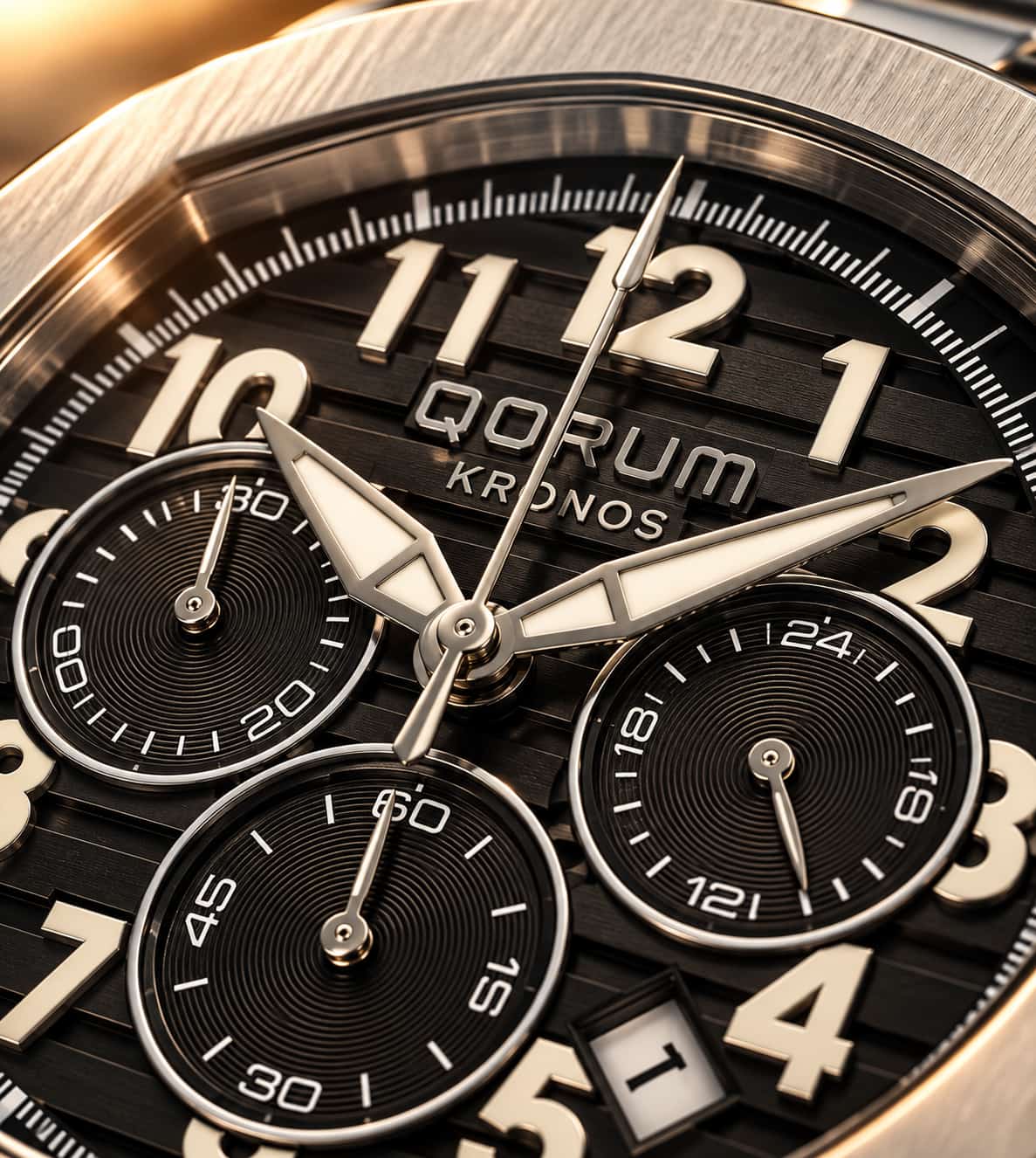 Close-up of a QORUM Kronos watch dial with black textured subdials, silver hands and markers, and a brushed metal case edge.