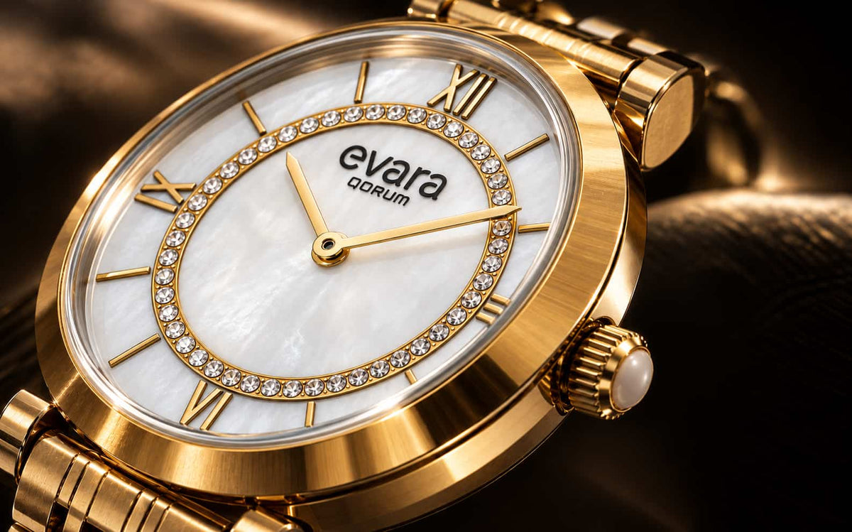 Close-up of a gold-tone wristwatch with a white mother-of-pearl dial, crystal rhinestone ring around the hour markers, and gold hands. The brand name 'evara QORUM' is visible on the dial.
