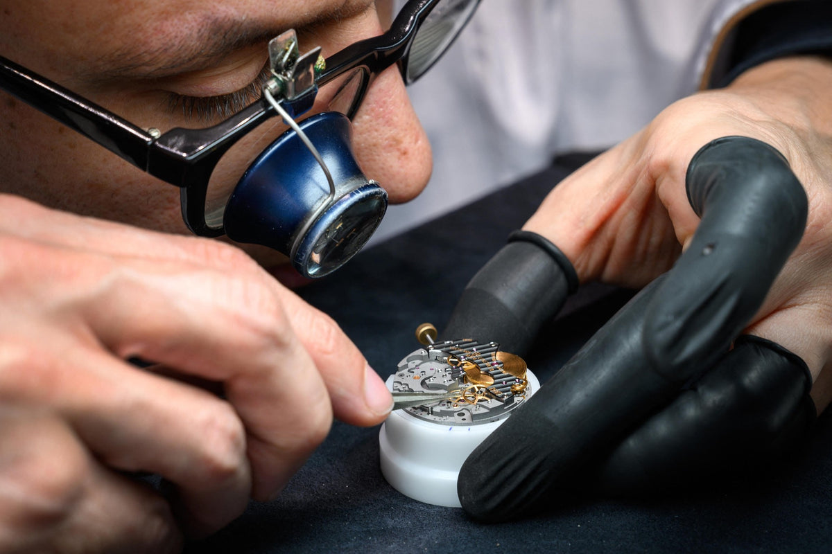 Close-up of a watchmaker wearing black gloves and a magnifying loupe, working on a small mechanical watch component mounted in a white holder.