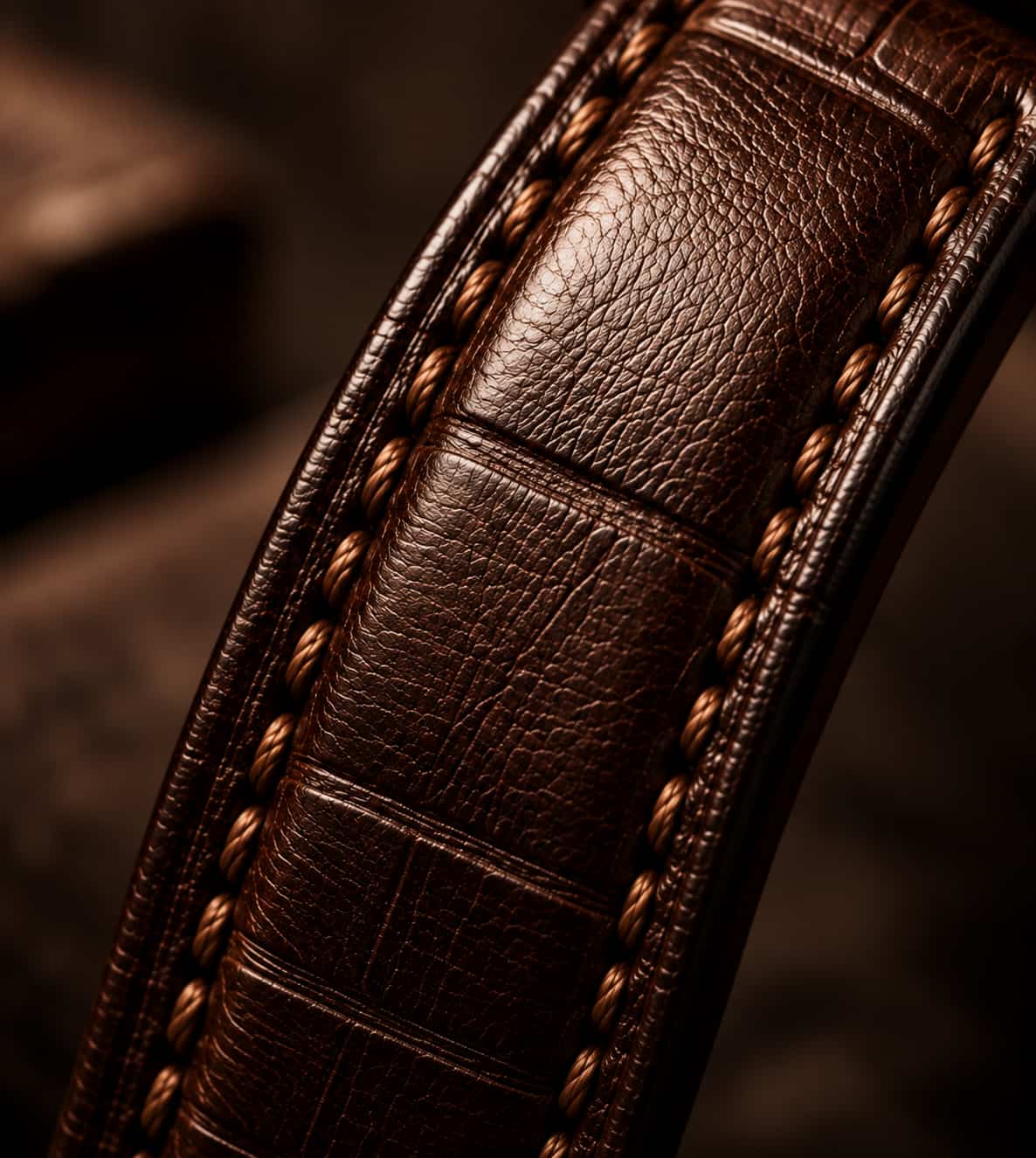 Close-up of a dark brown leather strap with crocodile-embossed texture and braided stitching, shown in warm, moody lighting.
