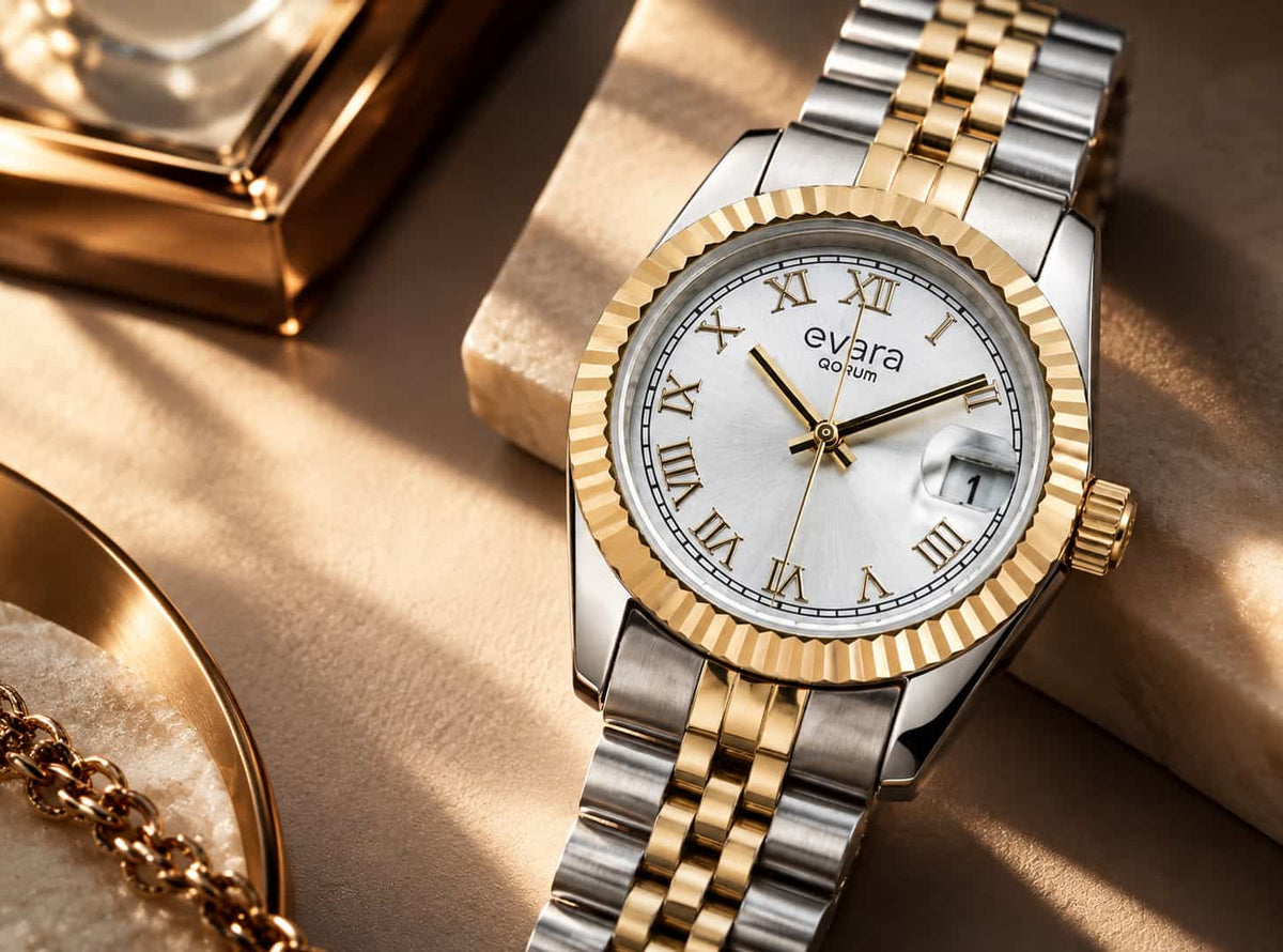 A close-up of an Evara Gold wristwatch with a two-tone silver and gold metal bracelet, a gold fluted bezel, and a white dial with Roman numerals, resting on a gold surface.