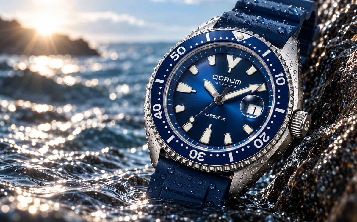 A QORUM Reef S automatic watch with a blue dial and blue rubber strap, resting on wet rocks in the ocean with waves and a bright sun in the background.