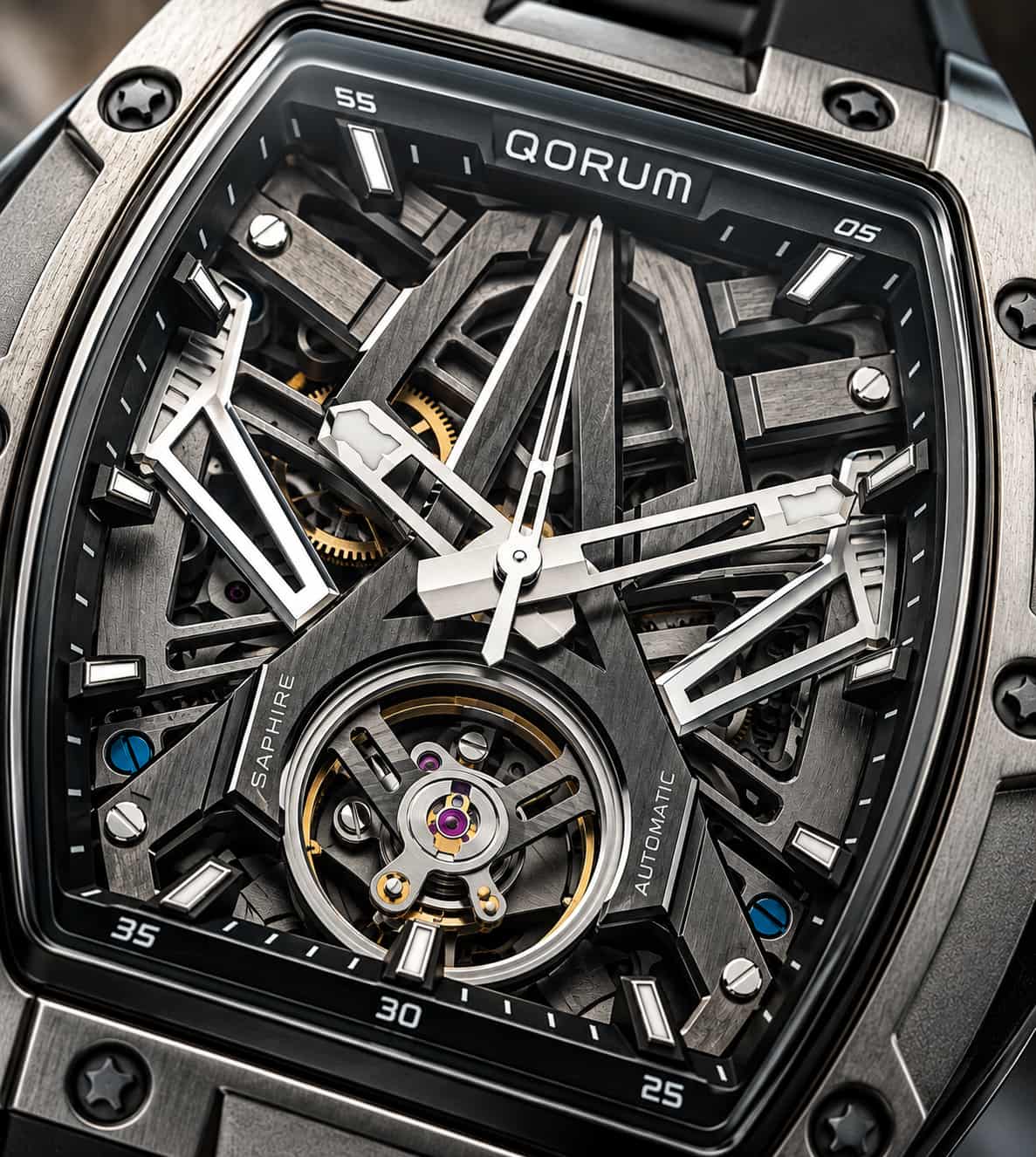 Close-up of a QORUM automatic skeleton watch with a visible tourbillon, featuring a dark metallic case, black dial, silver hands and markers, and gold-toned internal details.