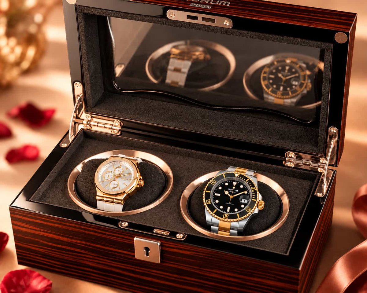 A luxury watch winder box with a dark wood grain exterior, black velvet interior, and two rotating watch holders displaying gold and silver-tone watches. The lid has a clear glass window showing two more watches inside, with rose petals scattered on a warm beige surface.