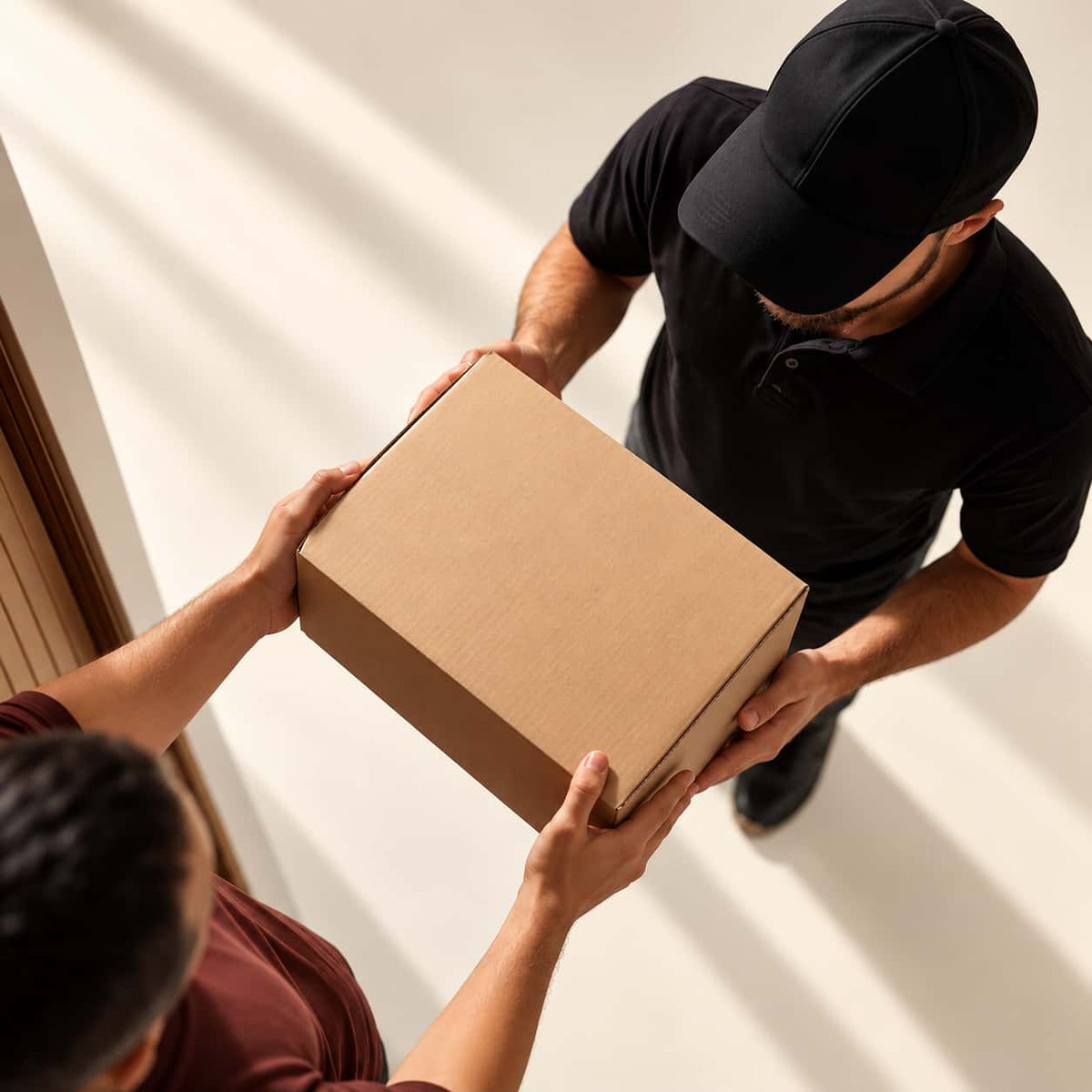 A cardboard shipping box being handed from a delivery person in a black uniform and cap to a receiver indoors.