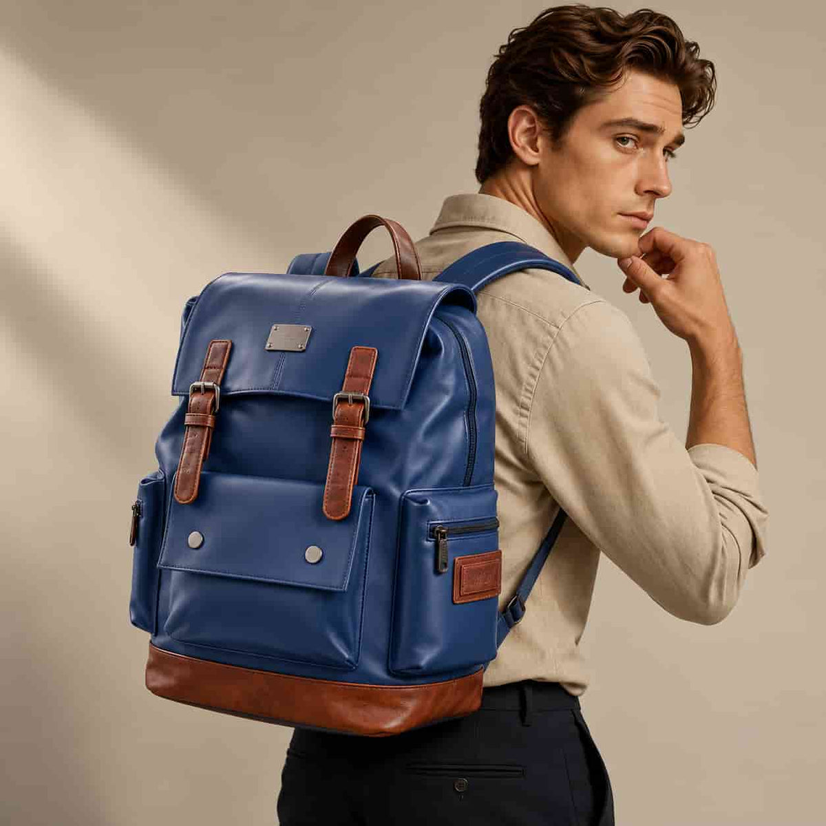 A man wearing a beige shirt and dark trousers carries a blue leather backpack with brown leather accents and silver-tone hardware against a warm beige studio background.