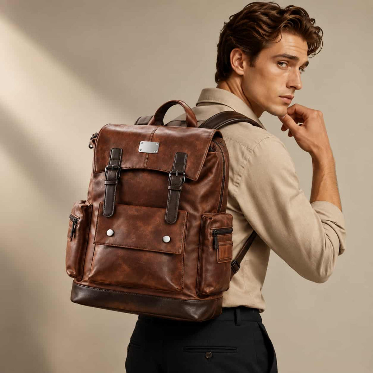 A man wearing a beige shirt and black trousers carries a brown distressed leather backpack with dark brown leather straps and silver-tone hardware, set against a warm beige background.