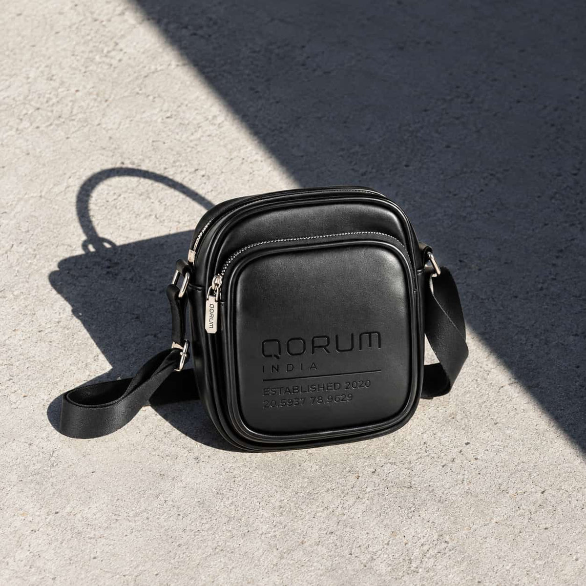 A black leather crossbody bag with silver hardware, featuring the brand name 'QORUM INDIA' embossed on the front, resting on a textured concrete surface under direct sunlight.