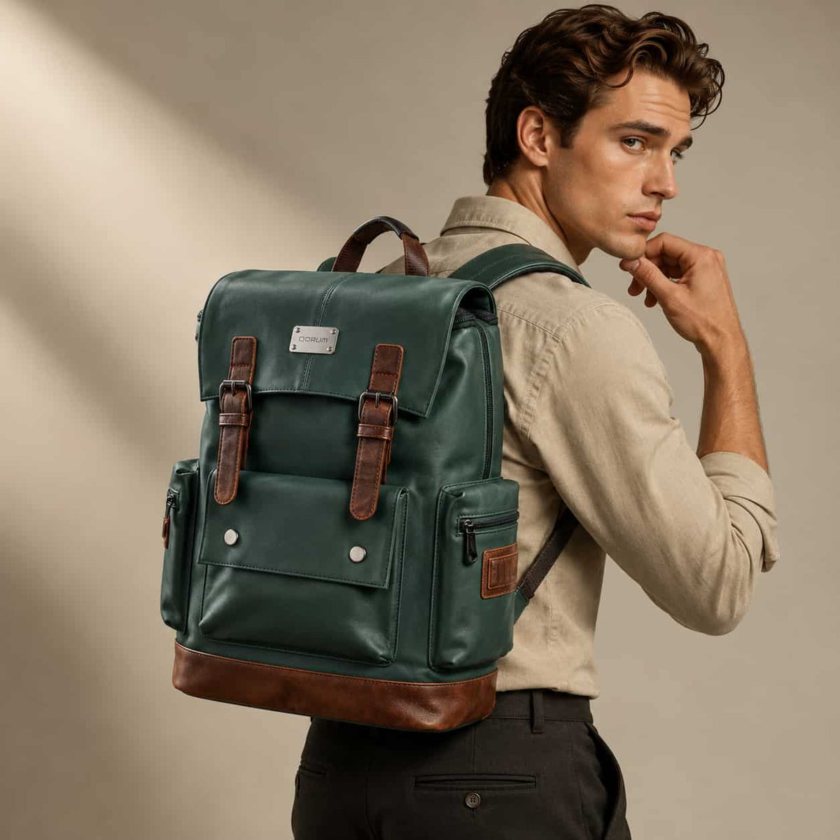 A man wearing a beige shirt and dark trousers carries a dark green leather backpack with brown leather accents and silver hardware, photographed against a warm beige studio background.