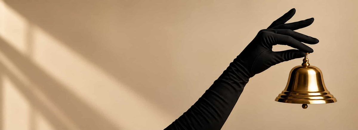 A black long-sleeve glove holding a polished gold bell against a warm beige background with soft window shadow patterns.