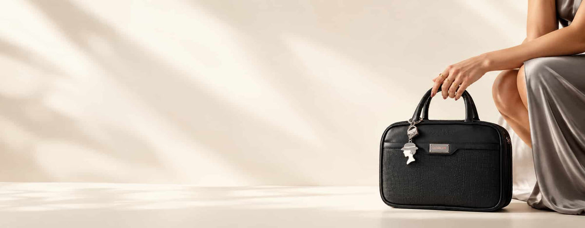 A black structured handbag with a top handle and hanging keychain, held by a person seated on the right against a warm beige background with soft shadow patterns.