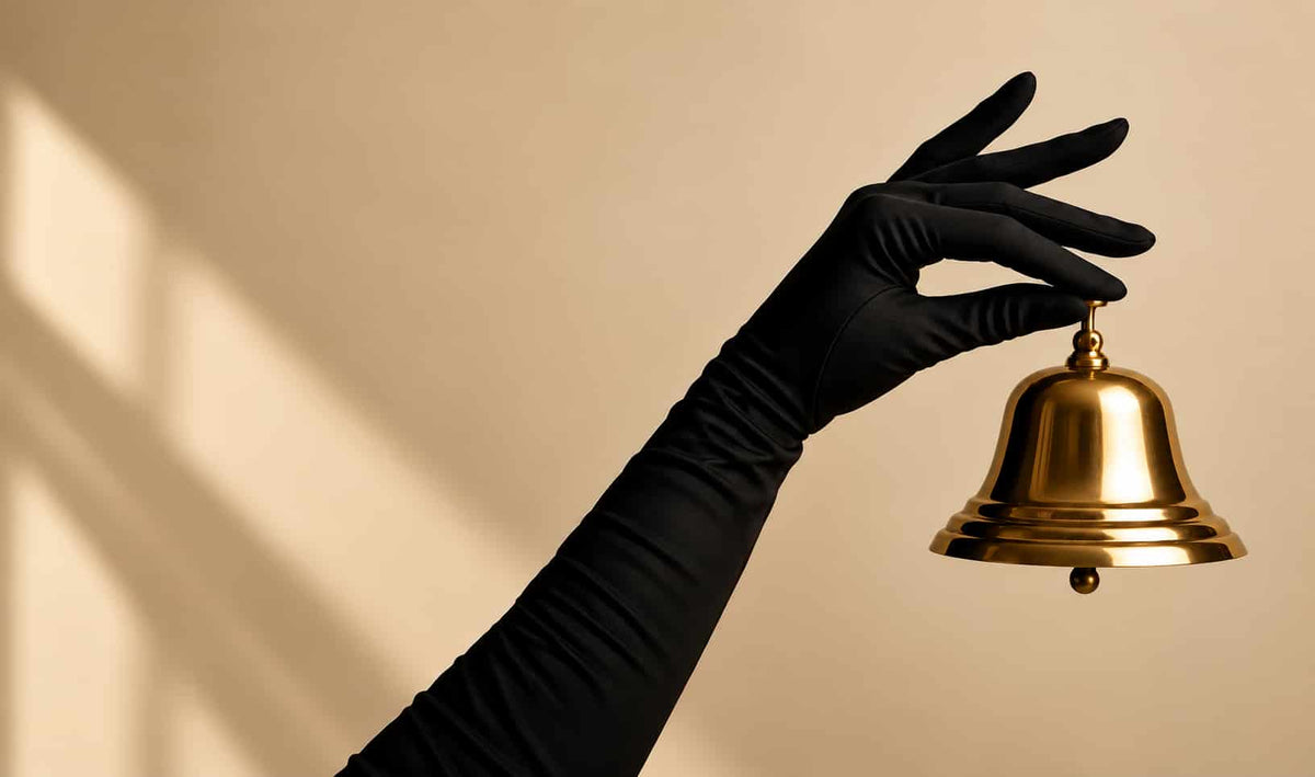 A black long glove or sleeve holding a small polished gold bell against a warm beige background with soft window-light shadows.