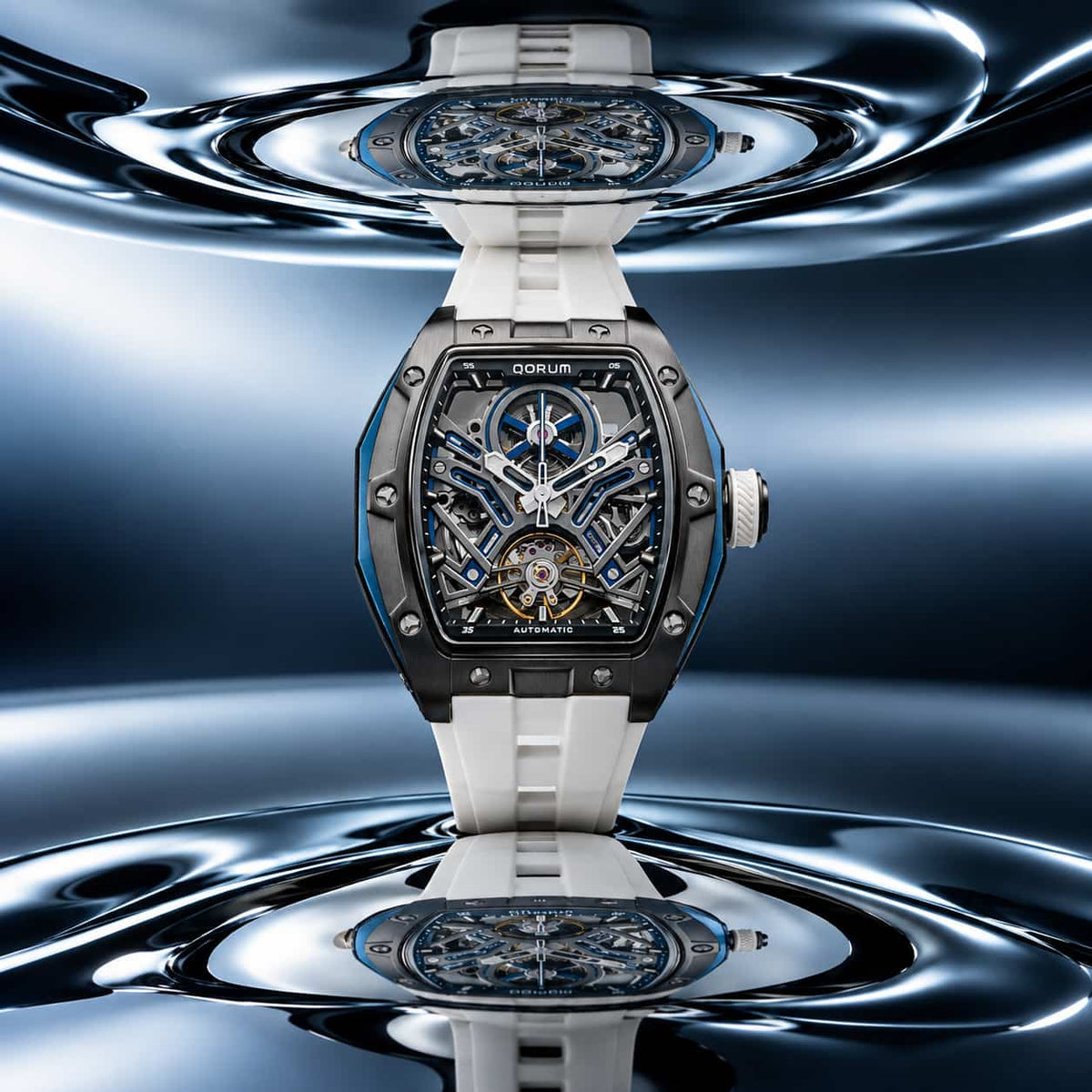 A luxury skeleton watch with a white rubber strap and a dark metallic case, displayed against a reflective blue and silver water-like background. The watch features a complex blue and gold mechanical dial.