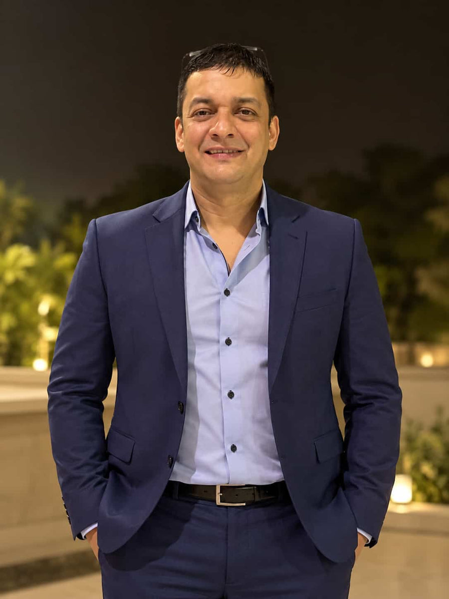 A man wearing a navy blue suit jacket and matching trousers over a light blue button-down shirt, standing outdoors at night with a blurred background of lights and foliage.