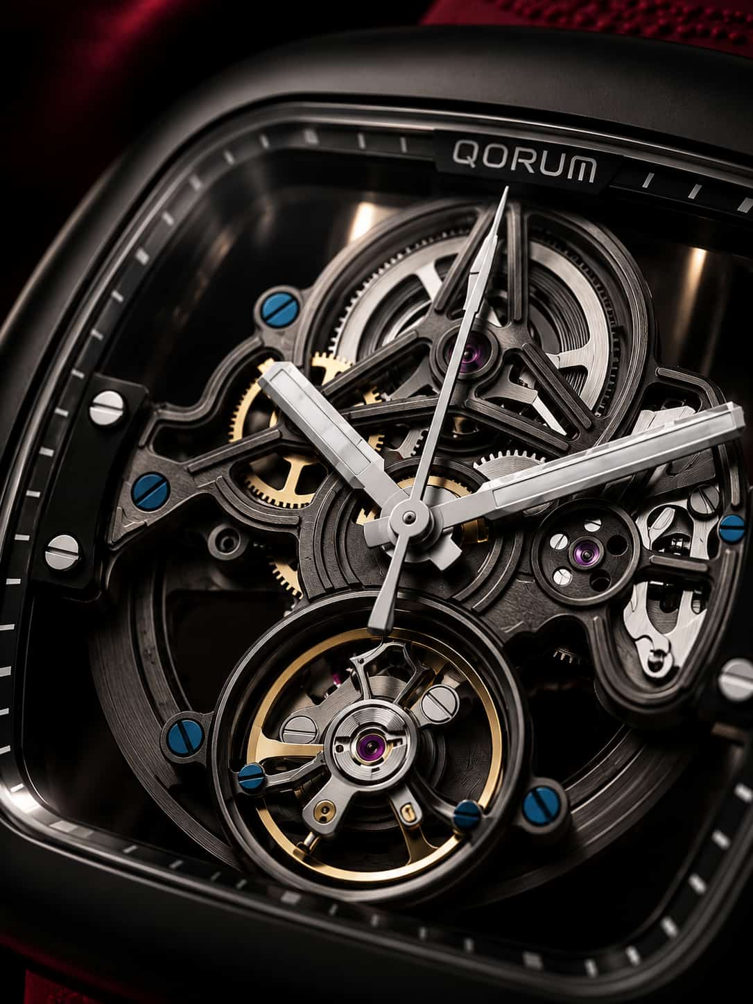 Close-up of a QORUM skeleton watch with a black case, silver hands, and a highly visible gold-toned tourbillon, showing intricate internal gears and blue jewel accents.
